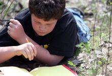 一个小男孩躺在草地上，手里拿着一支铅笔，低头看着一个红色的笔记本，里面写满了黄色的纸