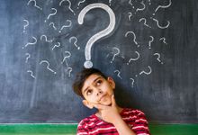 A young boy stands thinking in front of a blackboard covered with question marks.