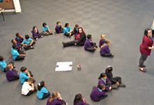 22 primary students and one teacher sitting in circle on the floor in assembly with two teachers standing on the side
