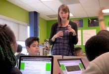 A female teacher is standing by and talking to four students at a table with their laptops open. 