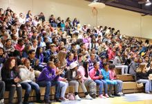 In the multipurpose room, a large group of middle school students fill one side of the bleachers, some clapping.