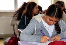 一个高中女生坐在一张紫色课桌旁的特写镜头。她在写，低头看着她的卷子。其他学生坐在她后面，有些模糊。