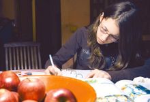 一个戴着眼镜的年轻女孩正坐在厨房桌子旁写作。桌上有一碗苹果，旁边是一本打开的课本，旁边是一本打开的用横格纸装订的活页夹。
