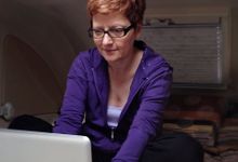 A red-headed woman in a purple sweatshirt with glasses is hunched over, sitting cross-legged, typing on her laptop. 