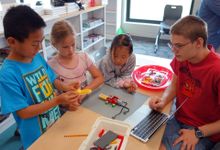 Four young students are sitting around a classroom table building a contraption out of legos and wires. It looks like it might be able to move prompted by technology.
