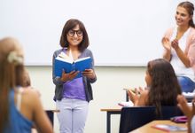 一个小女孩微笑着站在教室前面读书，她的老师和同学们都在为她鼓掌。