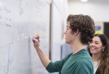 High school student writes math equation on board with teacher