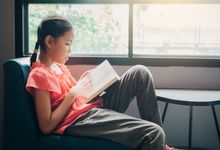 Middle-school aged girl reads a book while sitting in front of a window