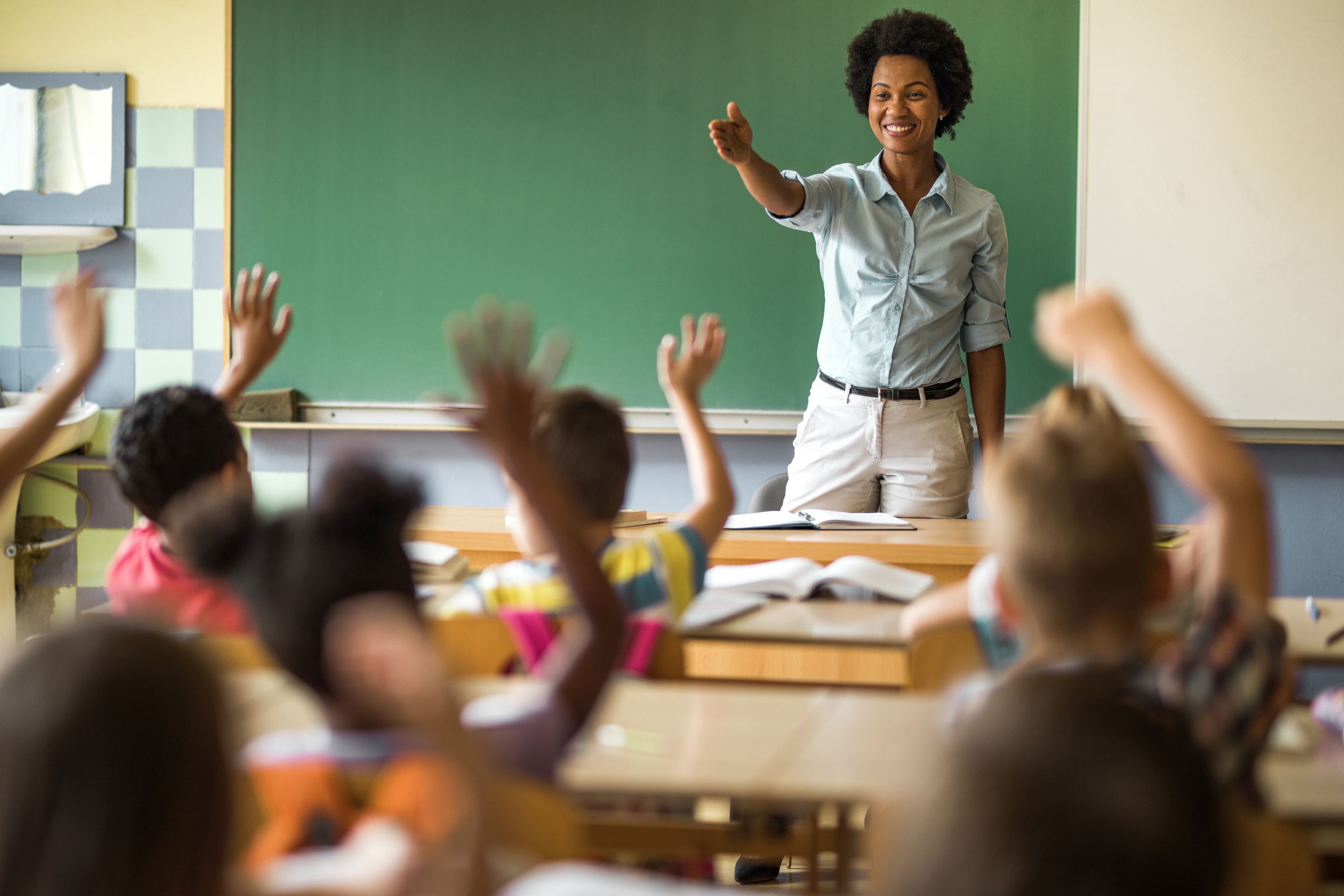  An elementary school teacher stands at the front of her classroom, smiling and pointing at something on the board while several students raise their hands.