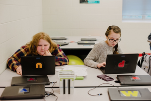 Two girls type on laptops in their classroom.