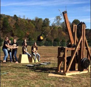 Teens are outside on a field catapulting pumpkins from a wooden contraption. 