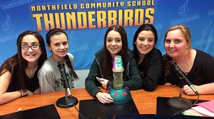 Five girls are sitting at a table with microphones behind a backdrop that says, "Northfield Community School Thunderbirds."