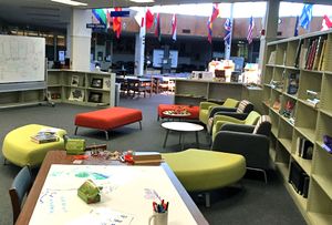 Large open space in the library with four brightly colored comfy chairs, large ottomans, desk, and white board