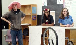 A tall teenage boy in a pink hat is standing at the front of the class with his hands on his hips. He's standing beside two teenage girls who are behind a desk turned into a podium. The three of them are smiling out to an audience.