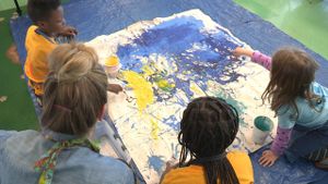 Three young students and a teacher are kneeling on a blue tarp in class pouring paint onto a large canvas.